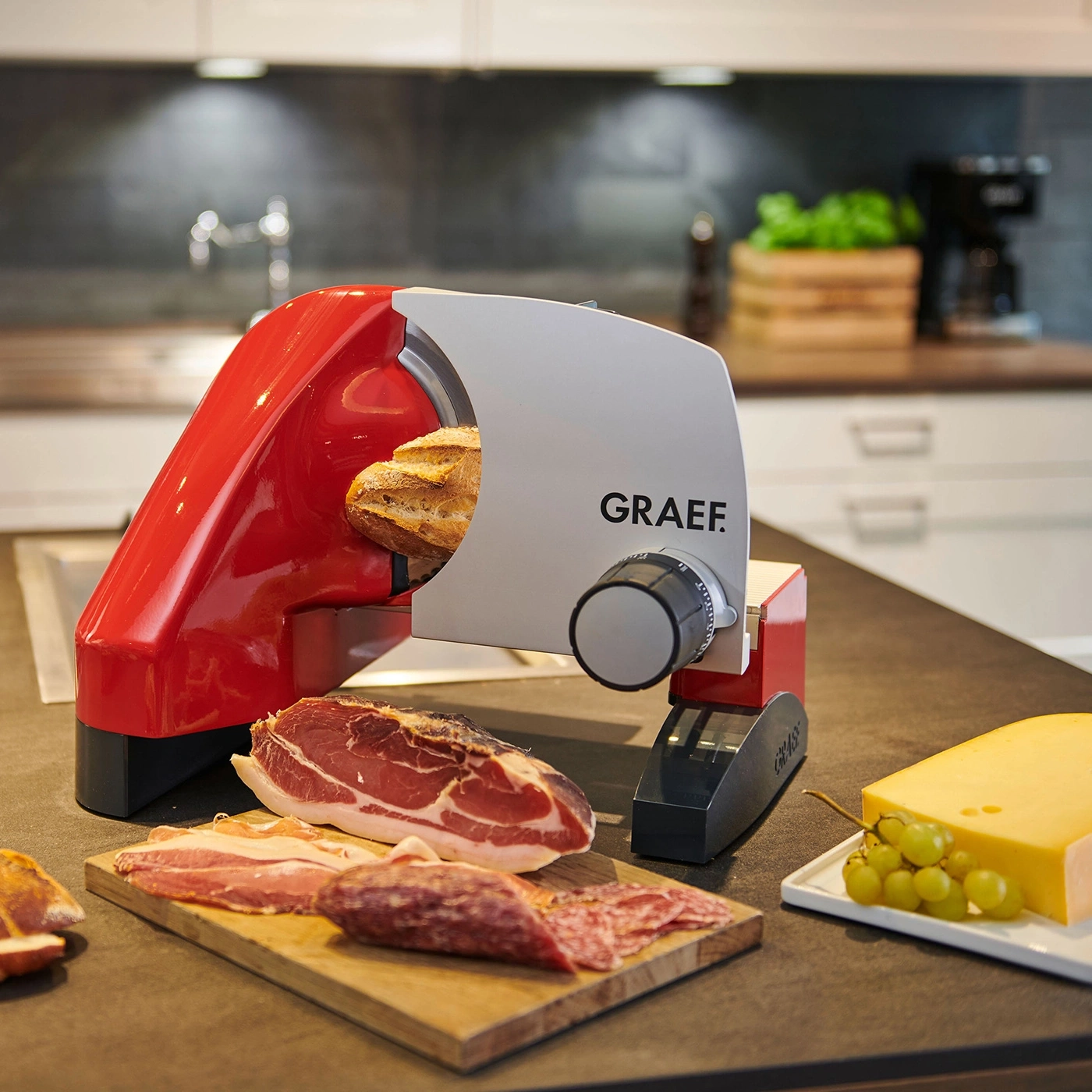 Affettatrice rossa con pane, carne, formaggio e uva su un bancone della cucina.