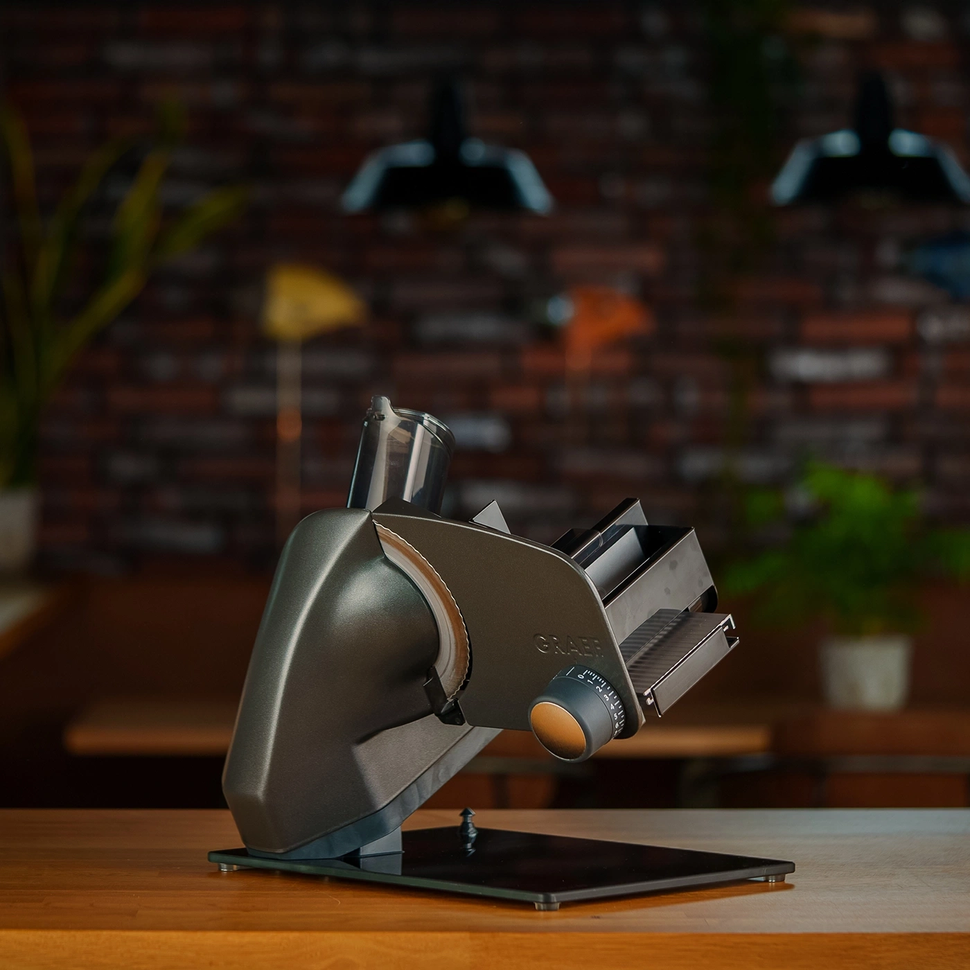 Une trancheuse Graef sur une table en bois, avec un mur de briques et des plantes en arrière-plan.