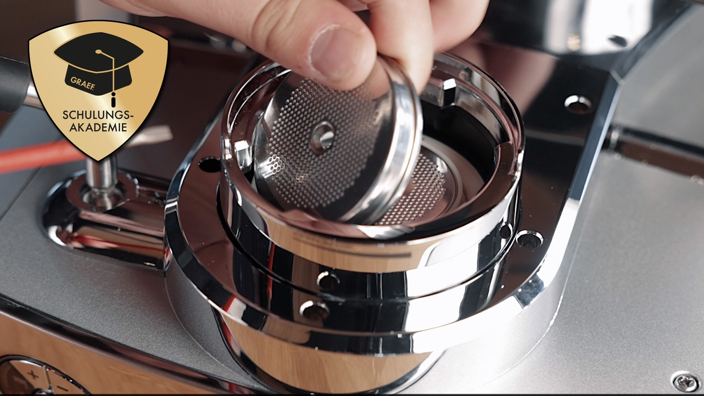 Hand holding portafilter insert in coffee machine, GRAEF Schulungs-Akademie logo