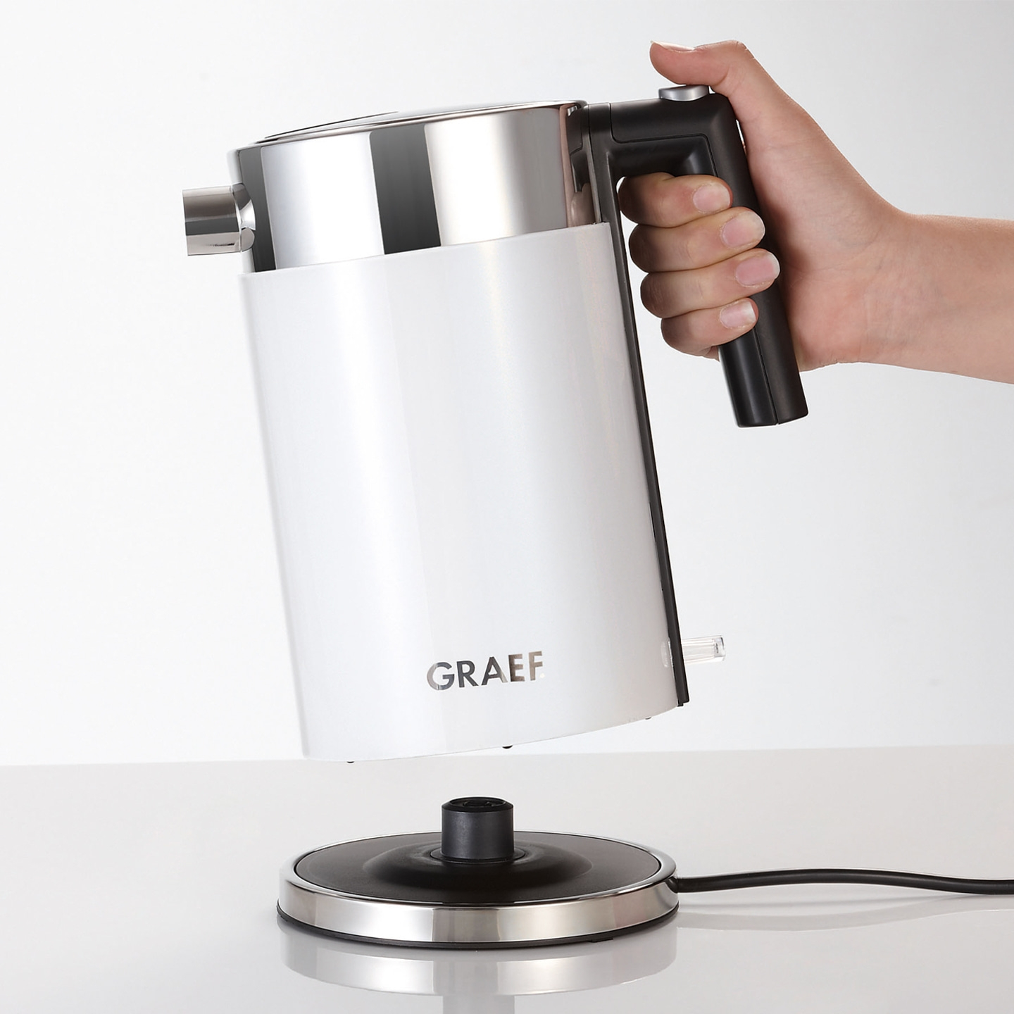 White kettle held by hand above base. The kettle has a black handle and silver accents.