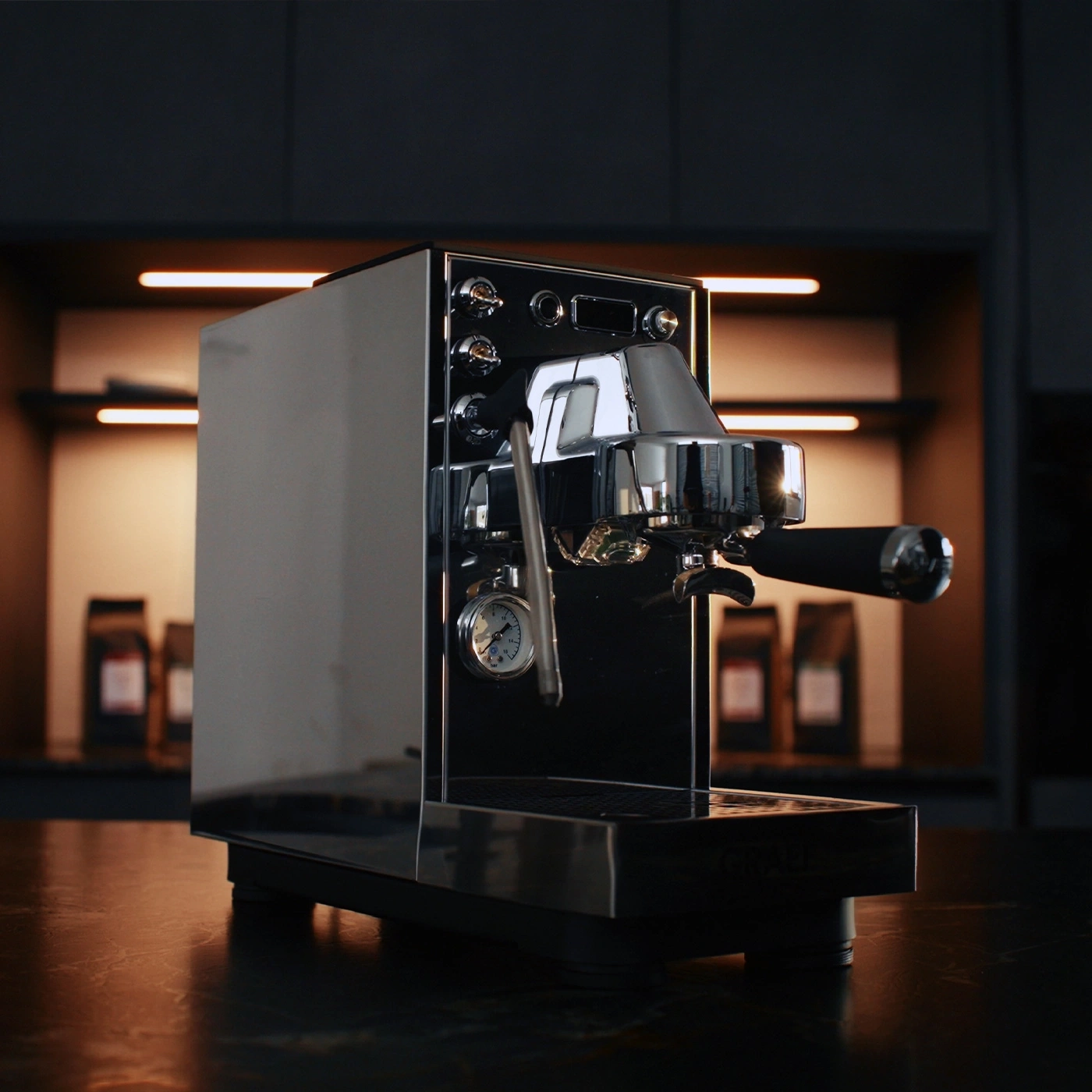 Une machine à expresso brillante est posée sur une table sombre, avec des paquets de café en arrière-plan.