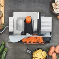 Trancheuse à légumes avec carottes, courgettes, pommes de terre et champignons sur fond gris.