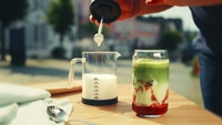 Person preparing drink, milk being poured, matcha drink, glass, wooden table, spoon