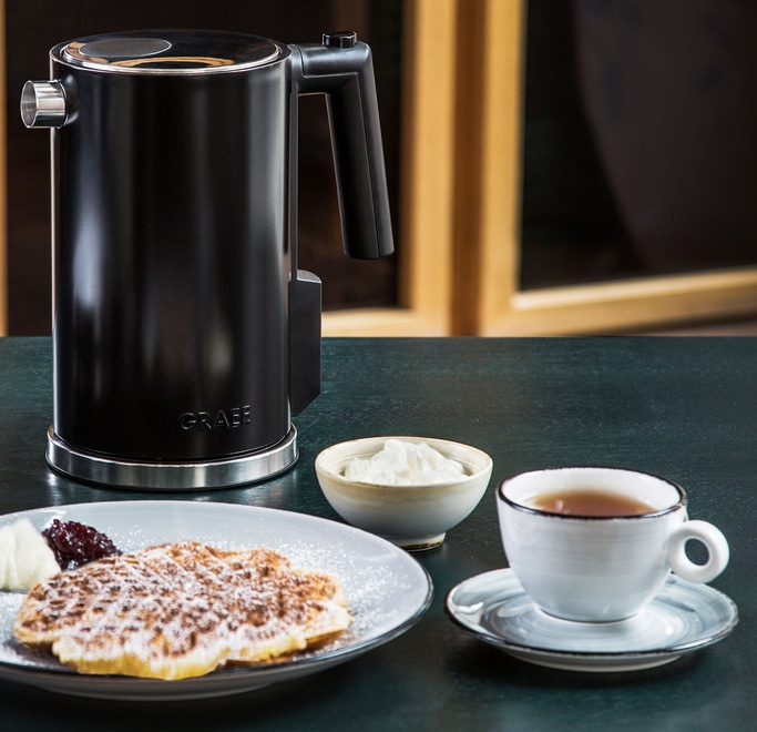 Black kettle, waffle, cream, jam, and tea on a table setting.