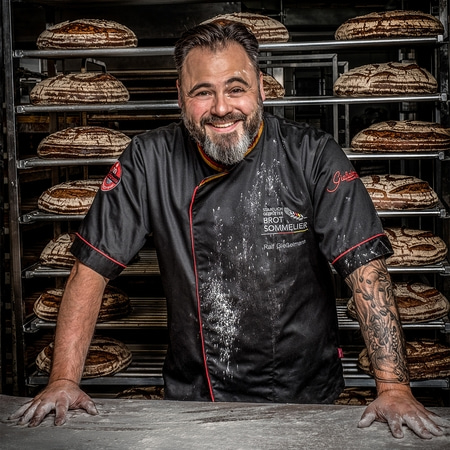 Bäcker mit Brot, lächelnd, in schwarzer Jacke, vor Brotregalen, Tattoo am Arm.