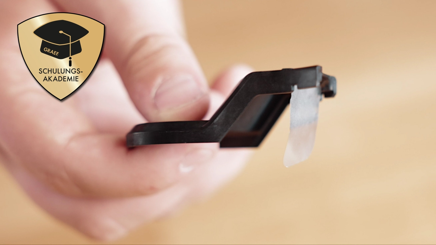 Objet noir avec logo tenu par une personne sur fond marron, avec un logo.