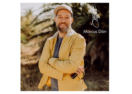 Homme barbu en veste jaune sourit, signature Marcus Dörr, fond flou