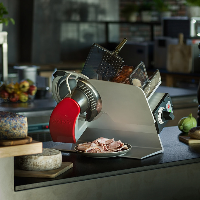 Meat slicer with ham, cheese, and vegetables on a countertop in a kitchen.