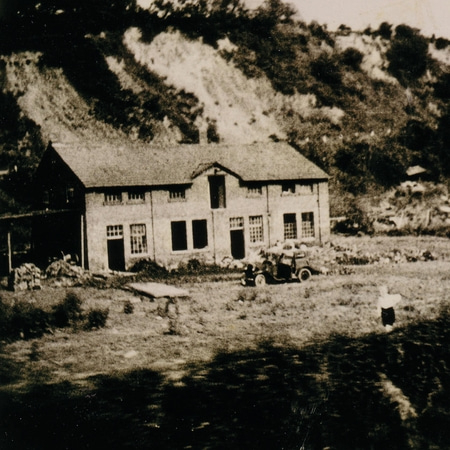 Vieux bâtiment avec voiture et personne devant une colline. Photo en noir et blanc.