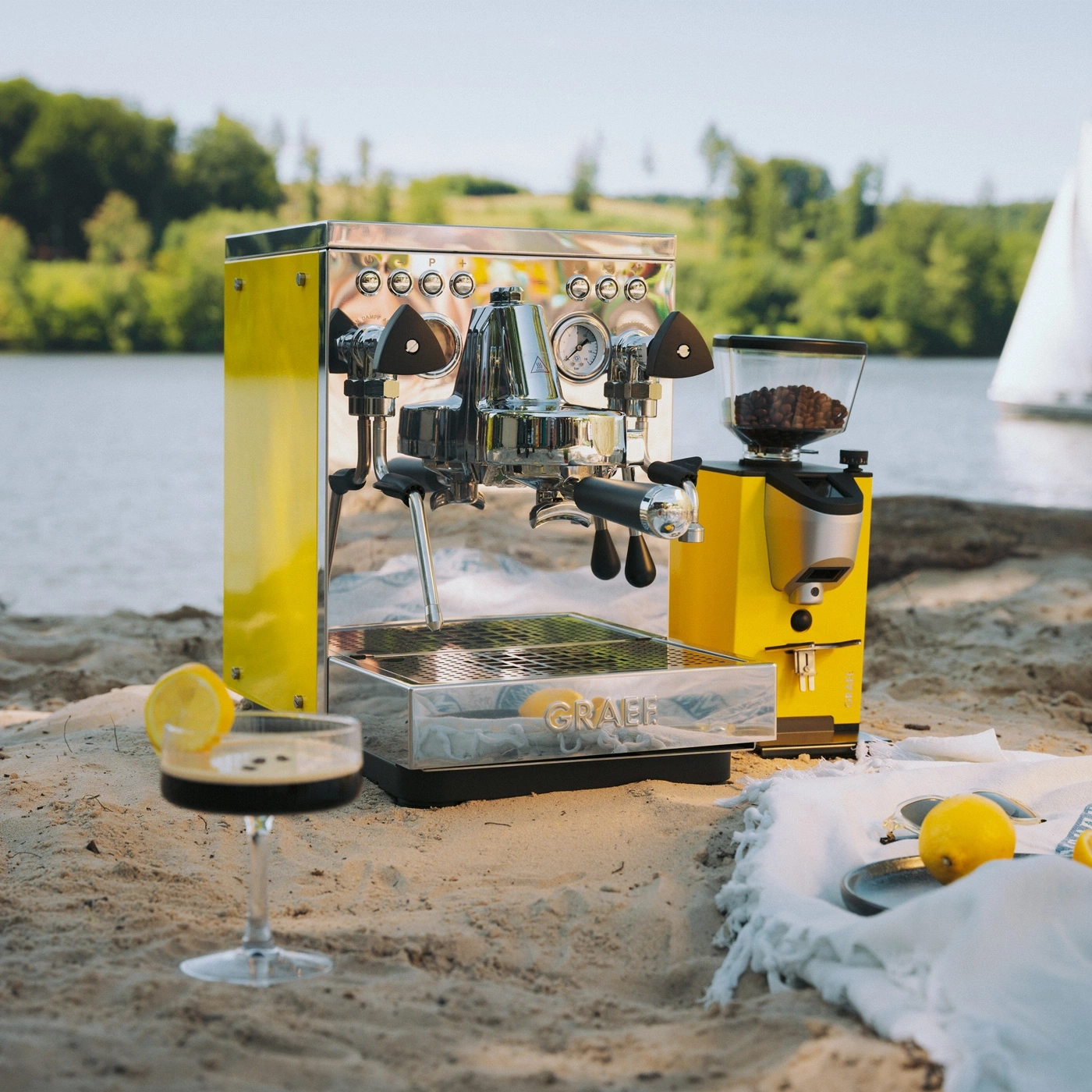 A yellow espresso machine and coffee grinder on the beach with a drink.