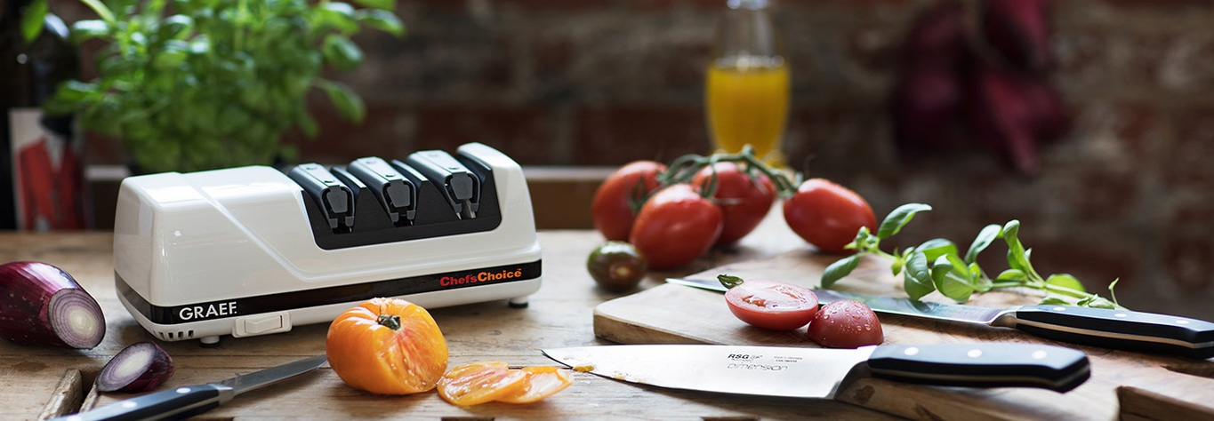 Küchenszene mit Messerschärfer, Tomaten, Zwiebeln und Kräutern auf Holz