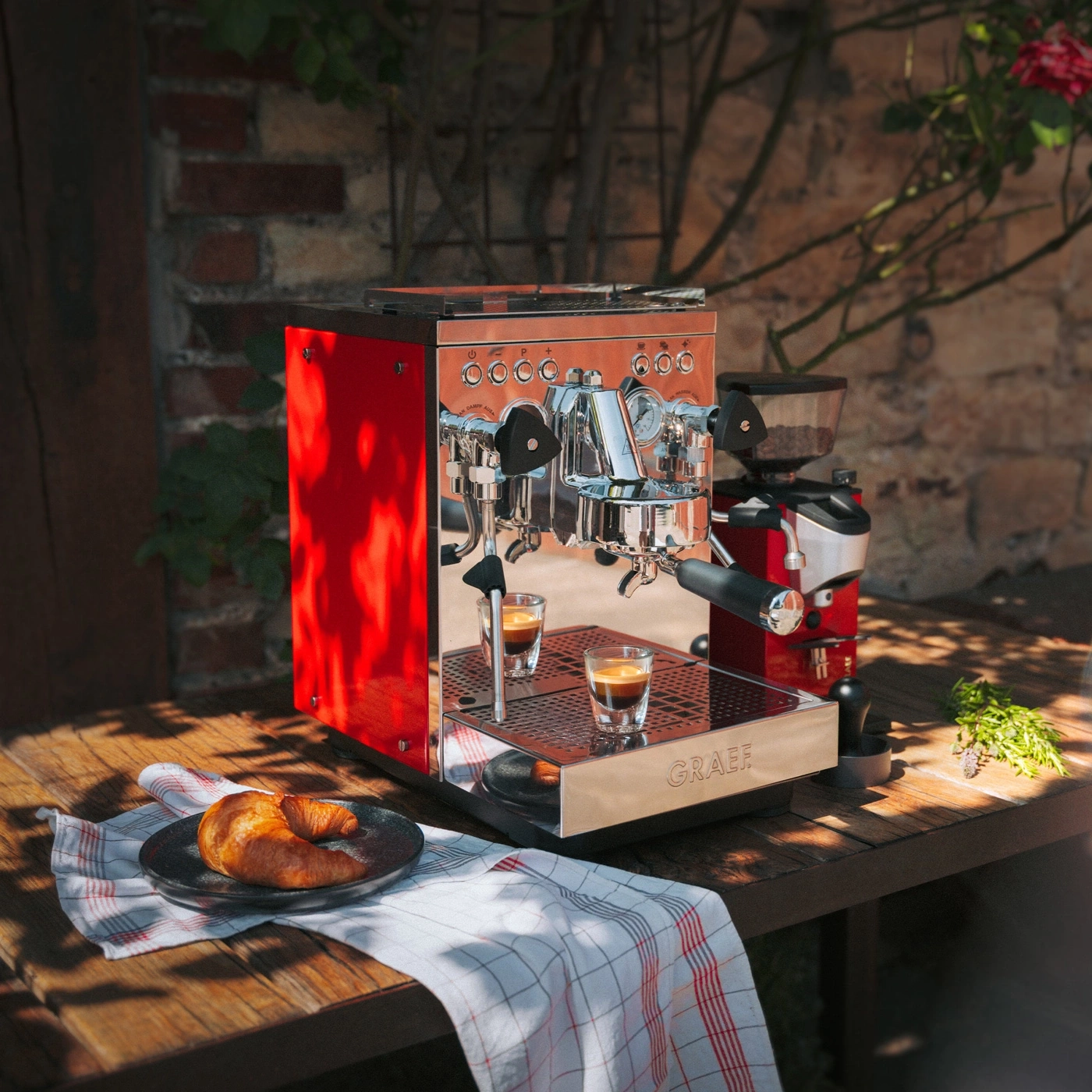 A red espresso machine with two shots of espresso, a croissant, and a coffee grinder outdoors.