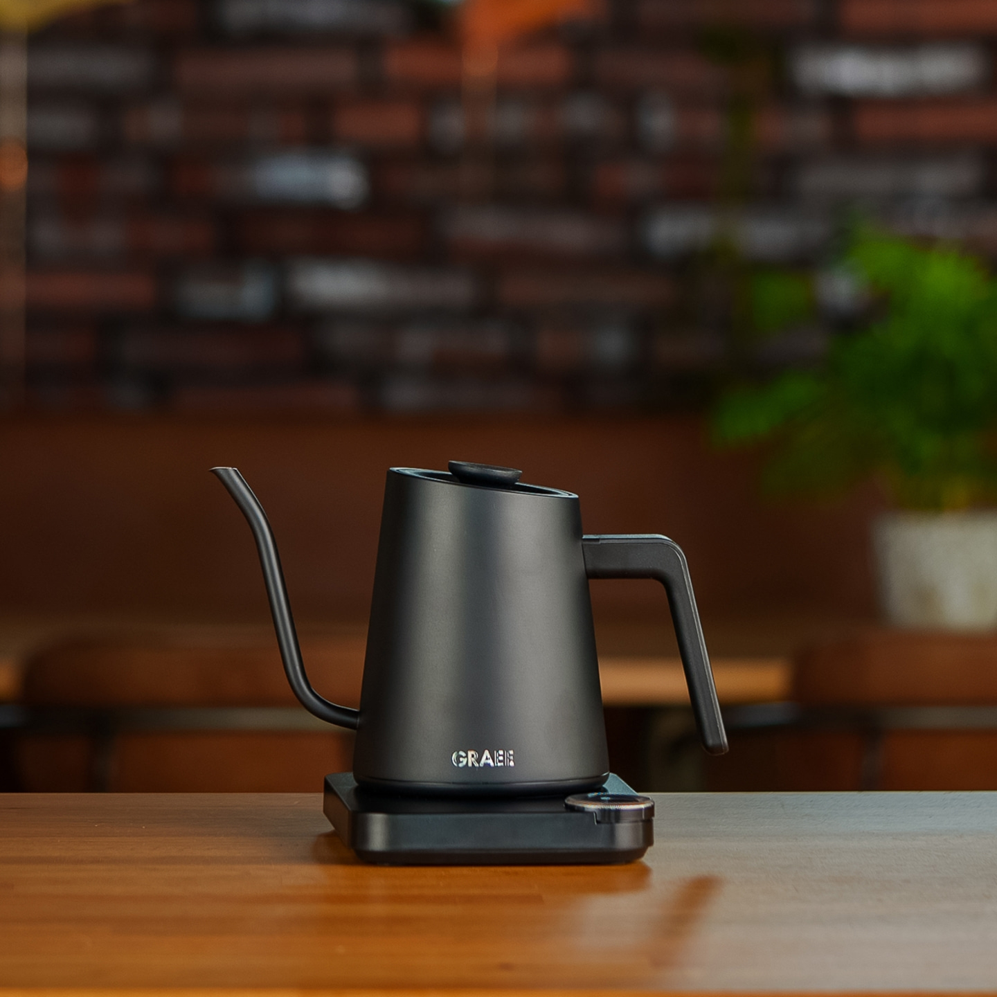 Black gooseneck kettle on wooden surface with brick and plant background.