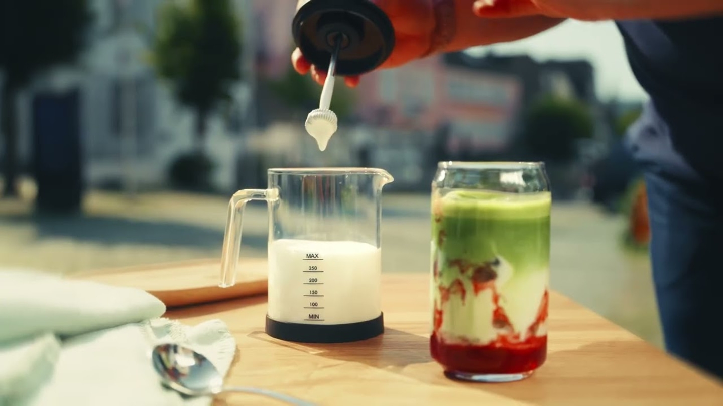 Person preparing drink, milk being poured, matcha drink, glass, wooden table, spoon
