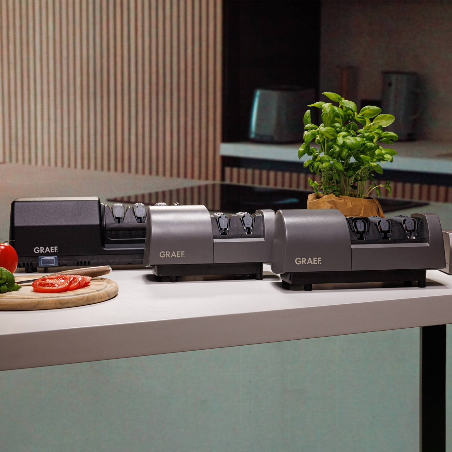 Three GRAEF knife sharpeners on a white surface with tomatoes and basil plant.