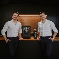 Two men pose with coffee machines. They are smiling, wearing white shirts.