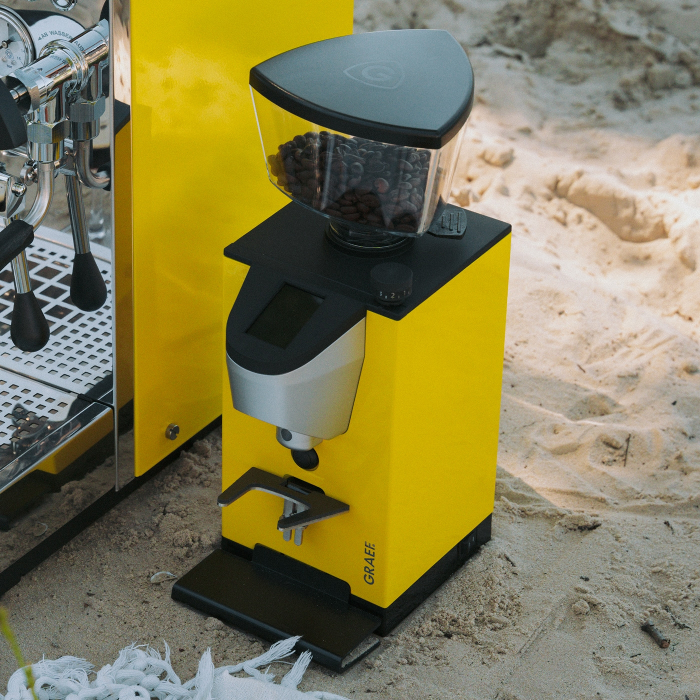 Un moulin à café jaune et une machine à expresso se tiennent sur la plage dans le sable.