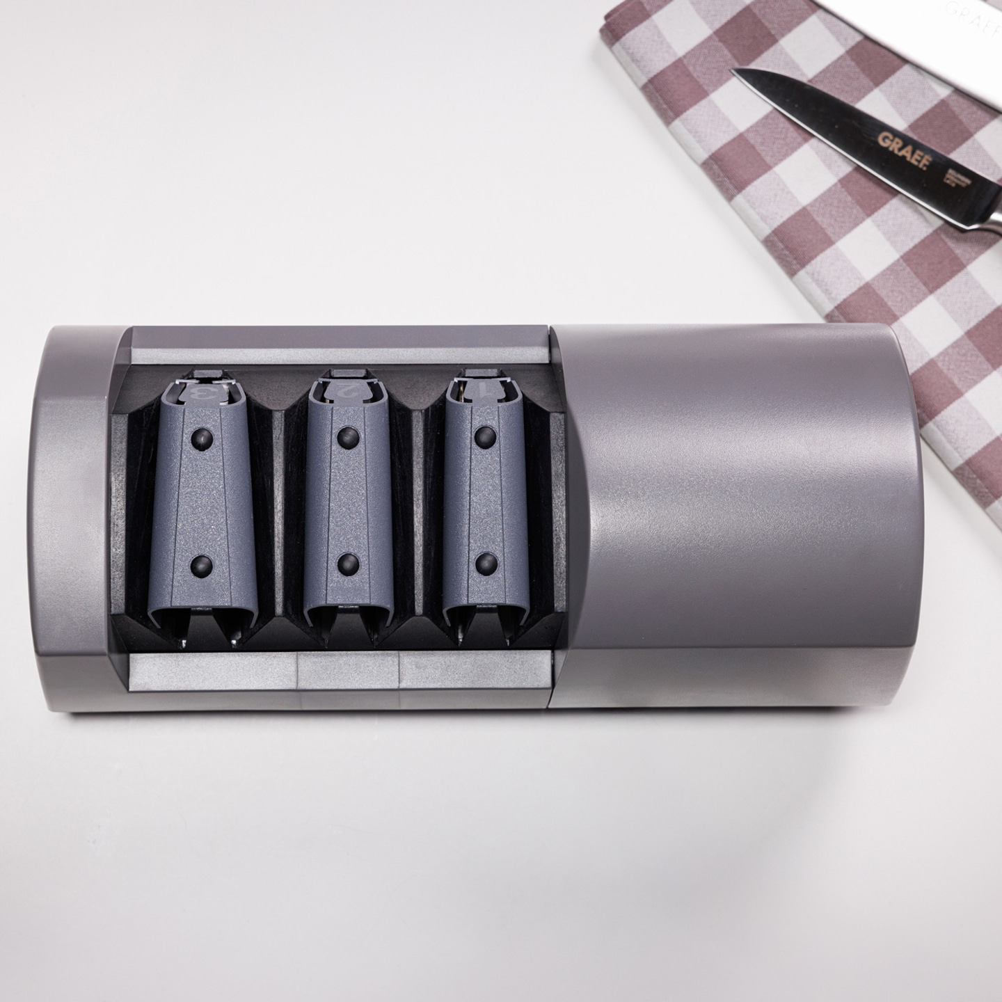 Affilacoltelli grigio con tre fessure e un coltello su un panno a scacchi.