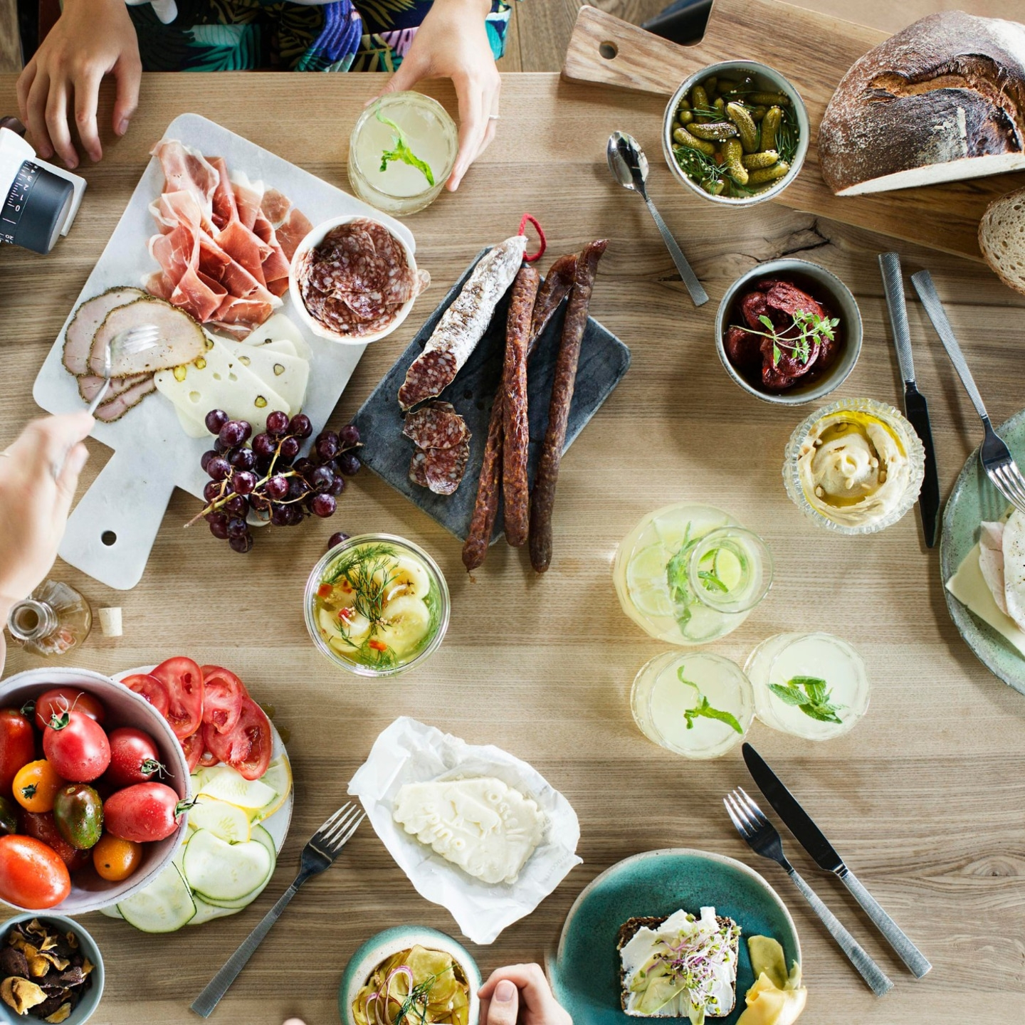 Tavolo di legno con cibo: salsicce, formaggio, verdure, bevande, pane, persone.