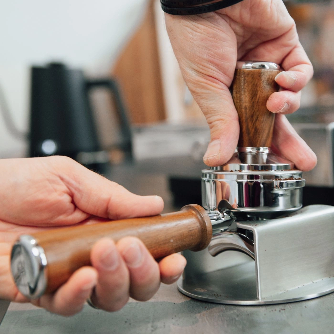 Mani che pressano la polvere di caffè con un tamper in un portafiltro, primo piano.