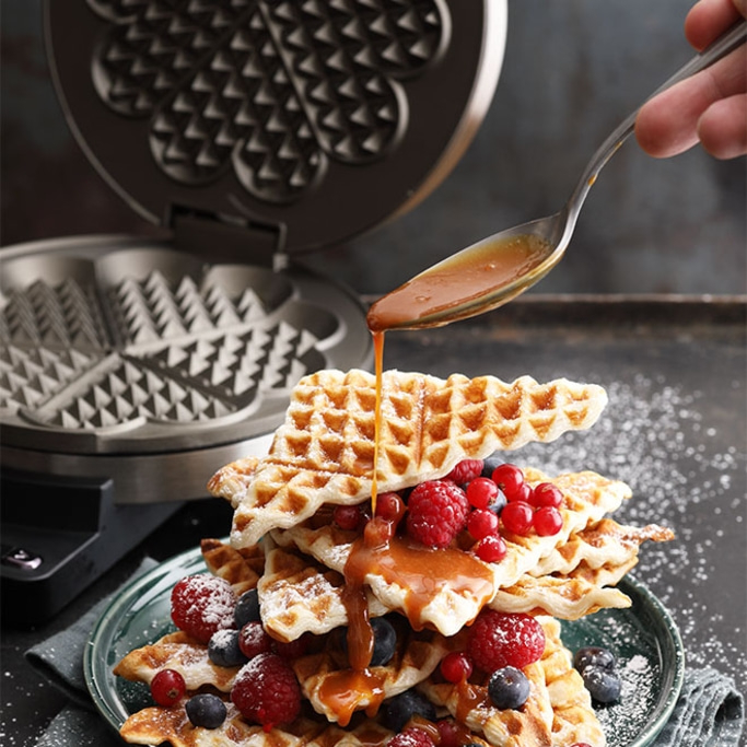 Waffles with berries and syrup being poured over them by a hand with a spoon.