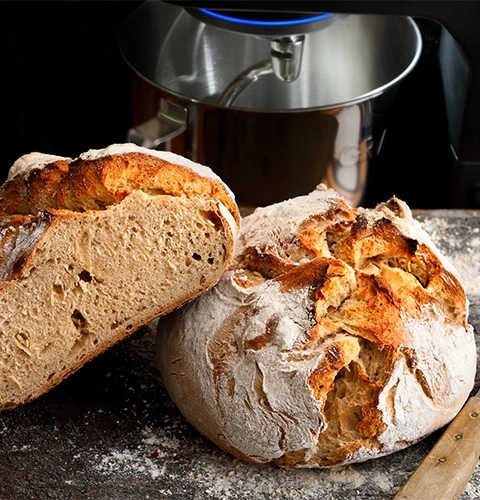 Pagnotta di pane affettata su tagliere di legno, con robot da cucina sullo sfondo