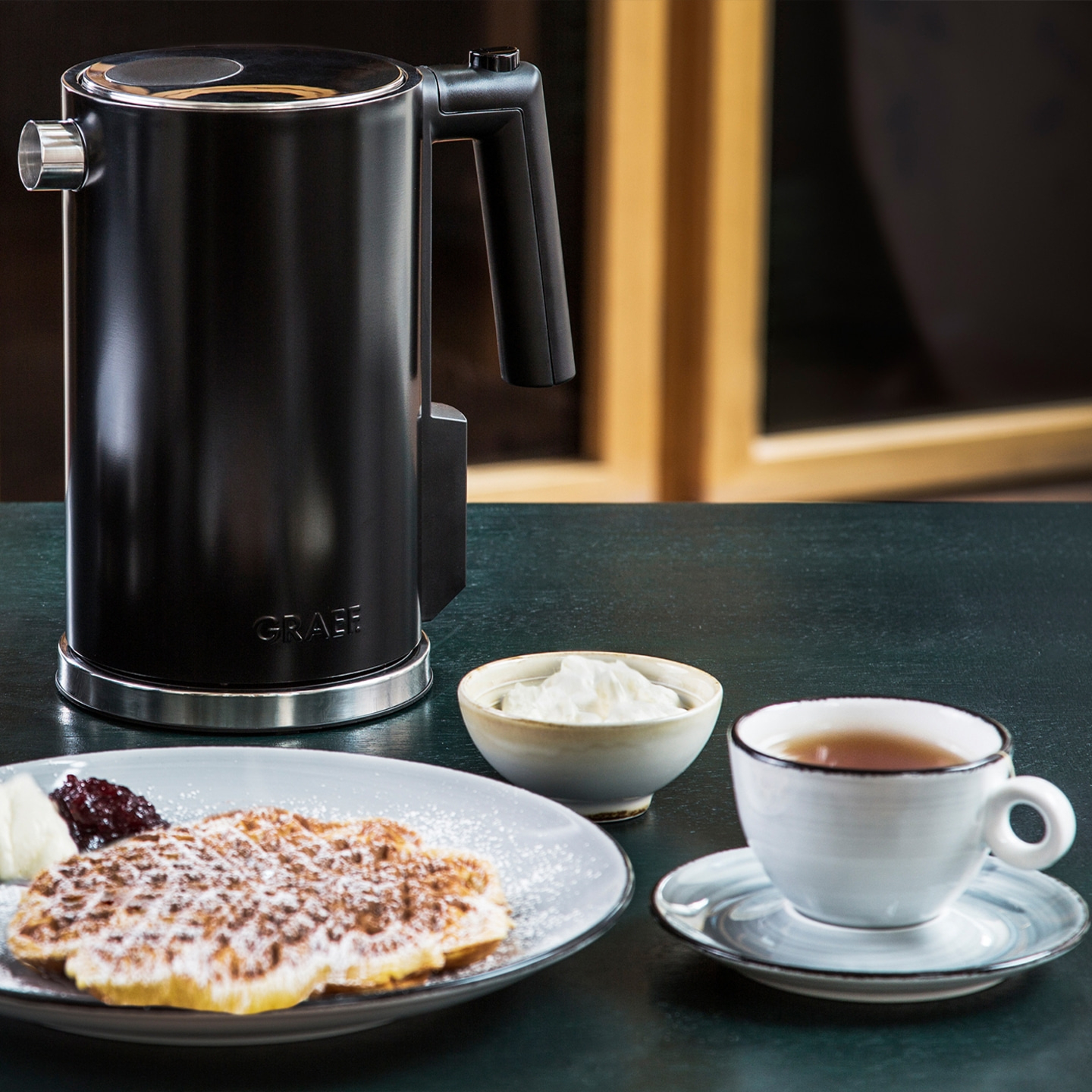 Black kettle, waffle, tea, cream, and jam on a table setting.