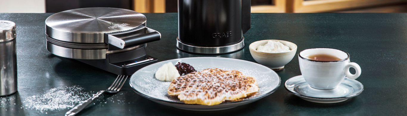 Waffle iron with waffle, cream, jam, and tea on a table setting.