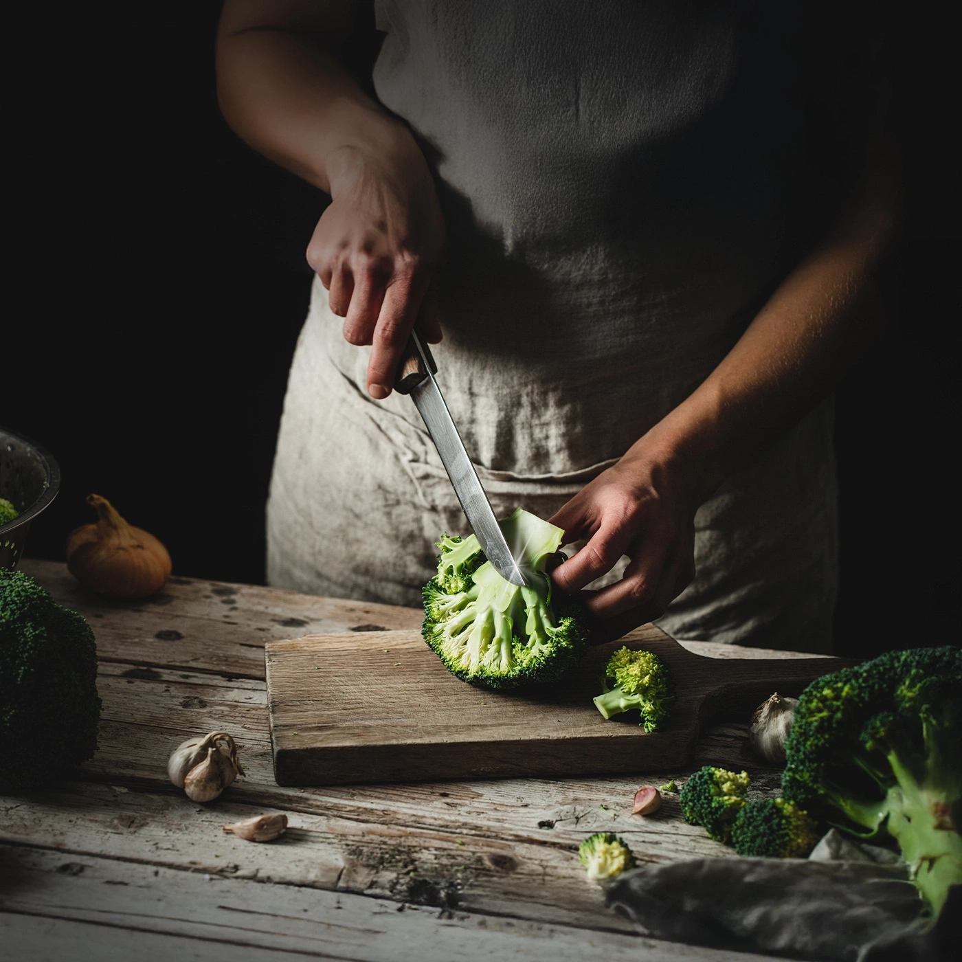 Una persona che taglia broccoli su un tavolo di legno con aglio e cipolle.