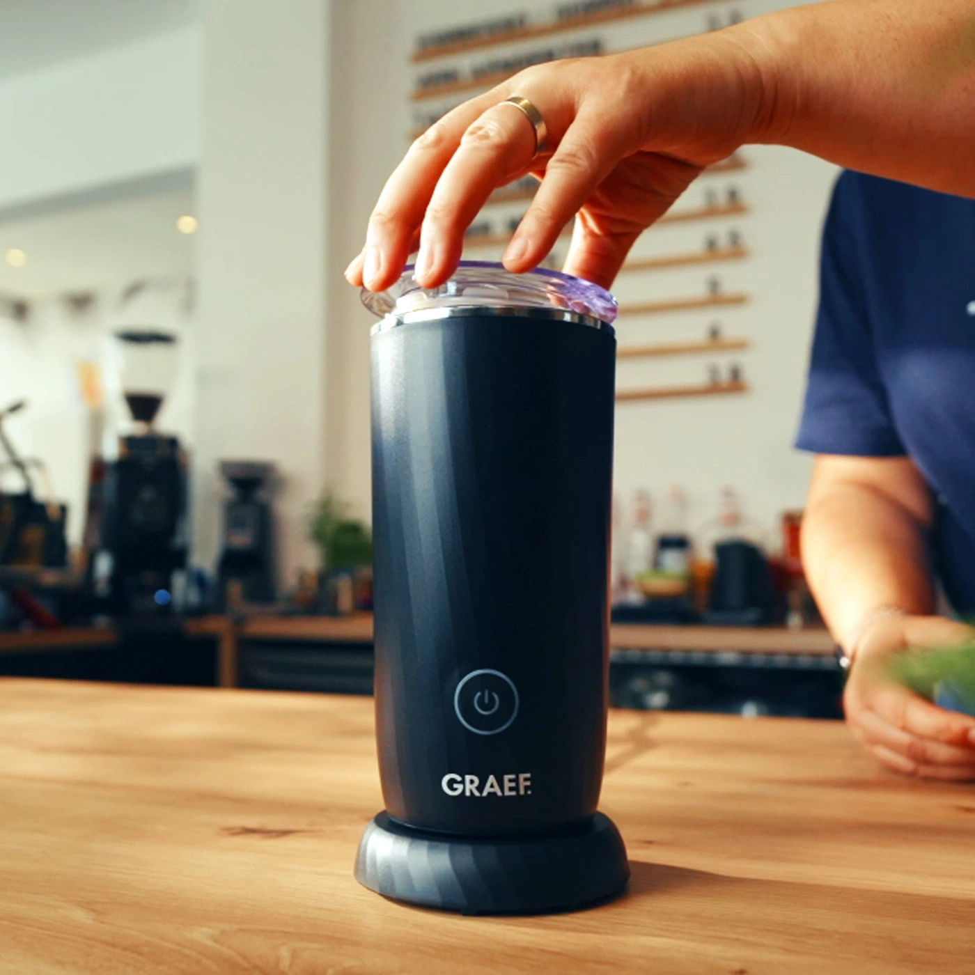 Une main ouvre un mousseur à lait noir avec le logo GRAEF.