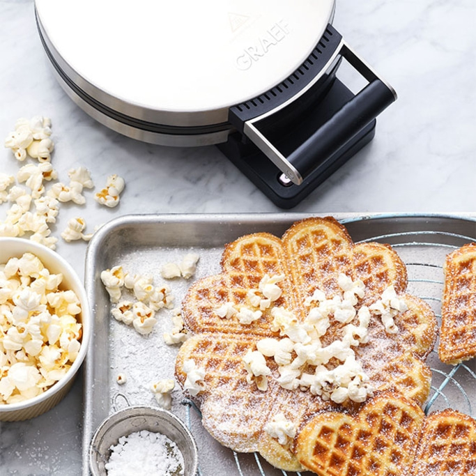 Waffle iron with waffles, popcorn, and powdered sugar on a tray