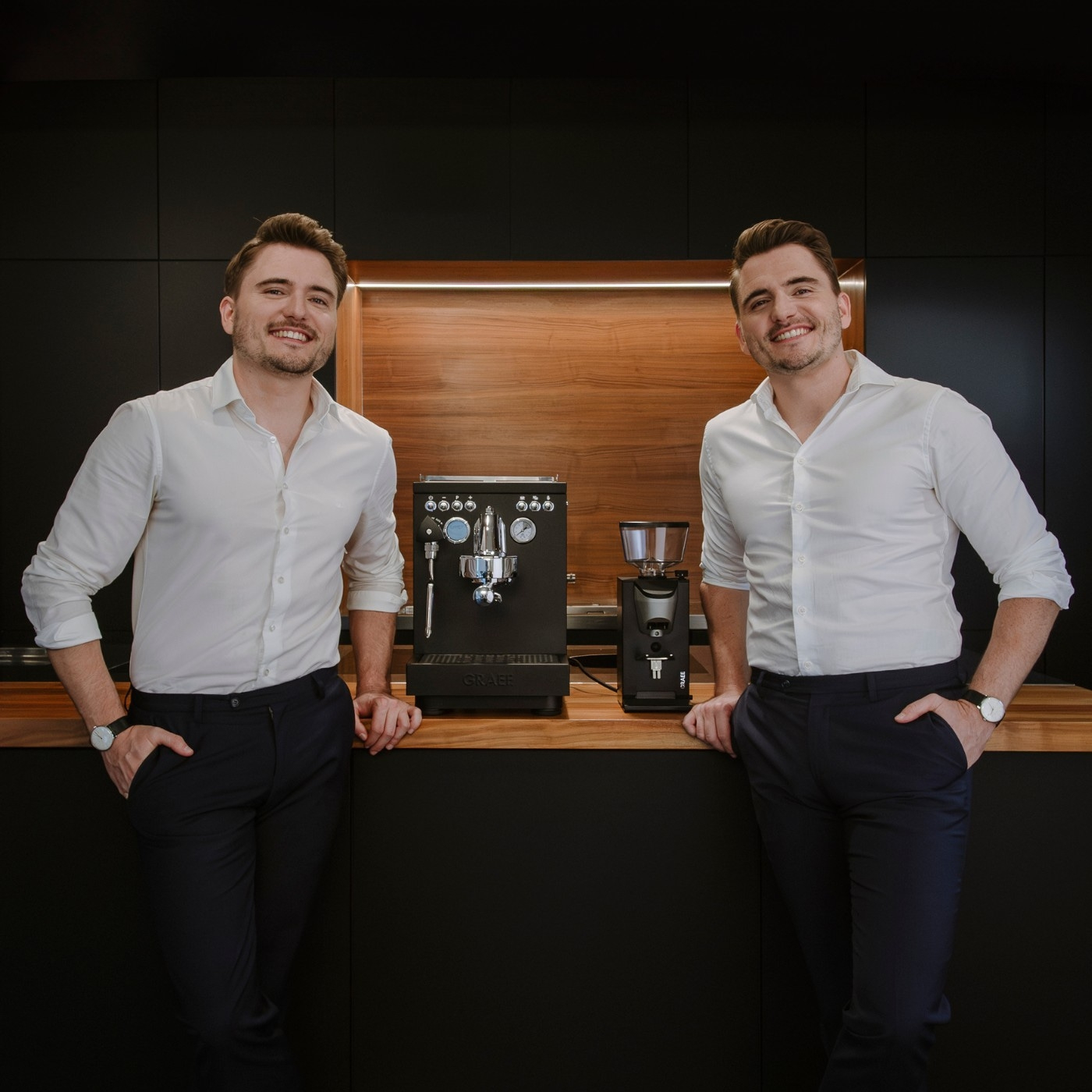 Two men pose with coffee machines. They are smiling, wearing white shirts.