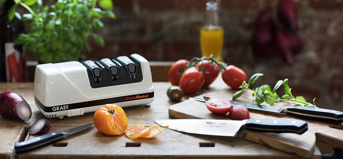 Scena di cucina con pomodori, coltelli, affilatore e verdure su superficie in legno