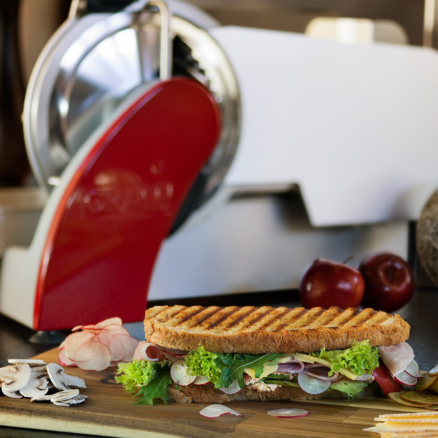 Sandwich with vegetables and meat, next to a slicer machine and apples