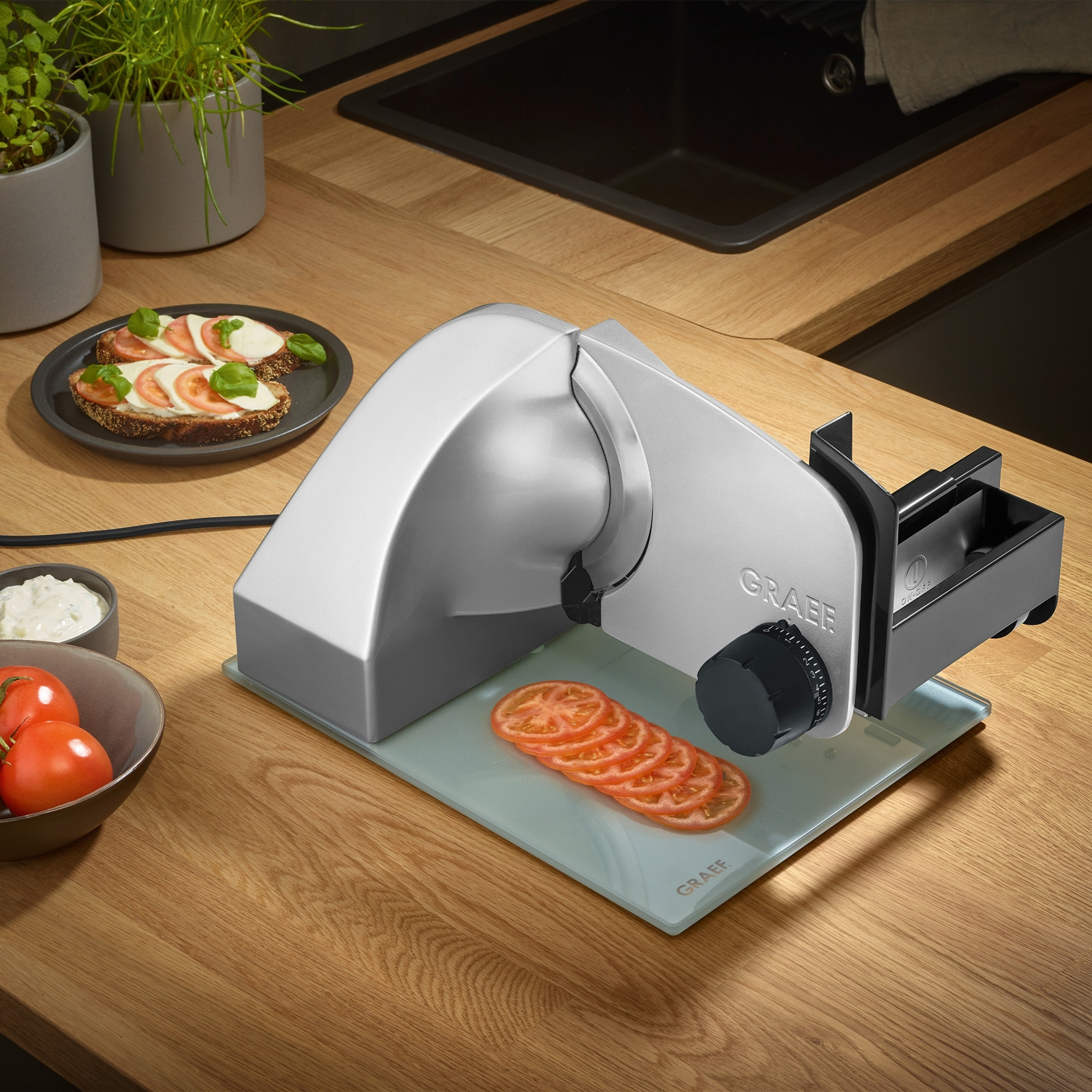 Fette di pomodoro su tagliere accanto ad affettatrice e cibo su piatto