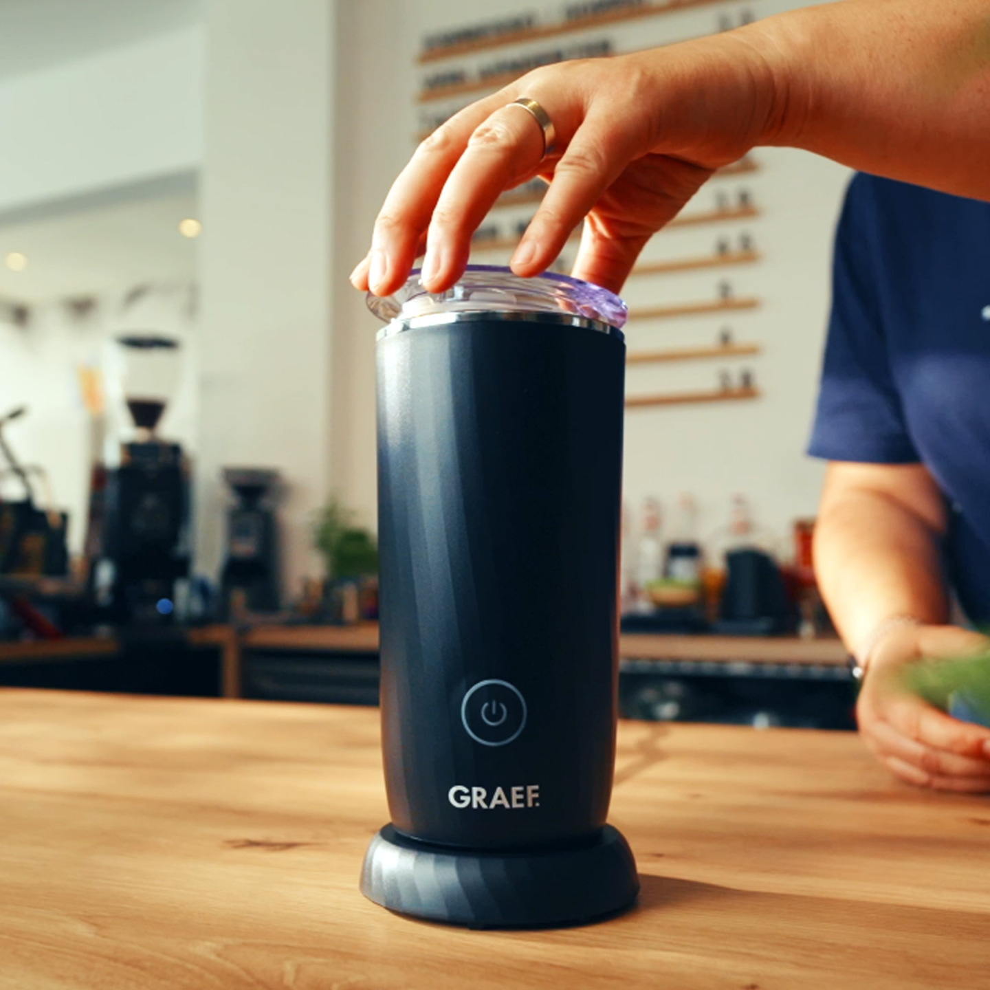 A hand opens a black milk frother with the GRAEF logo.